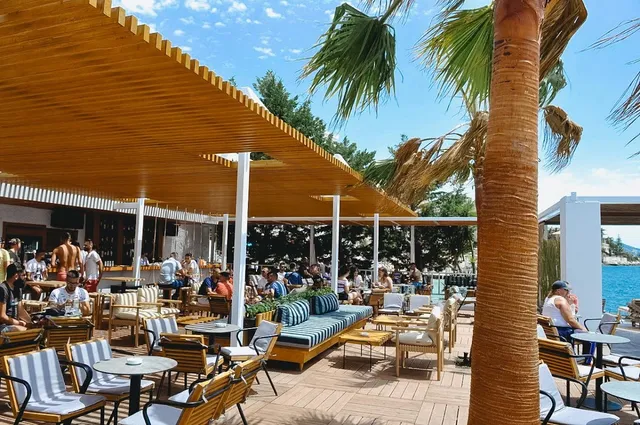 A shaded wooden terrace with blue striped sofas, palm trees and blue water to the side