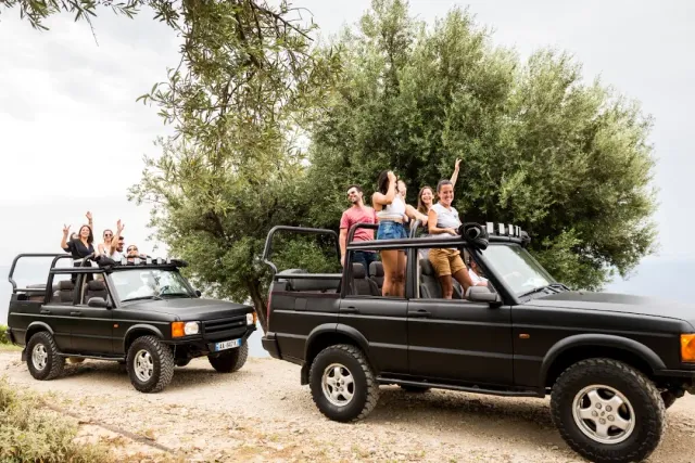 Two open black jeeps with passengers standing and waving, next to an olive tree