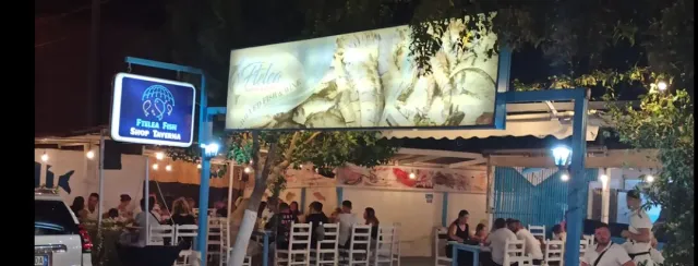 A taverna at night with lit signs, white chairs and diners under hanging bulbs