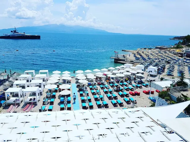 A view from above of a beach packed with white umbrellas and sunbeds, with a yacht in the bay