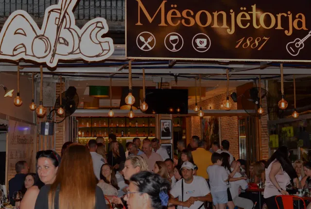 A busy venue at night with a lit sign, hanging bulbs and a crowd sitting outside
