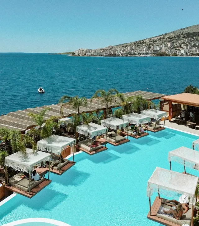 A turquoise pool with shaded sunbeds above the sea, and a town on the hill opposite