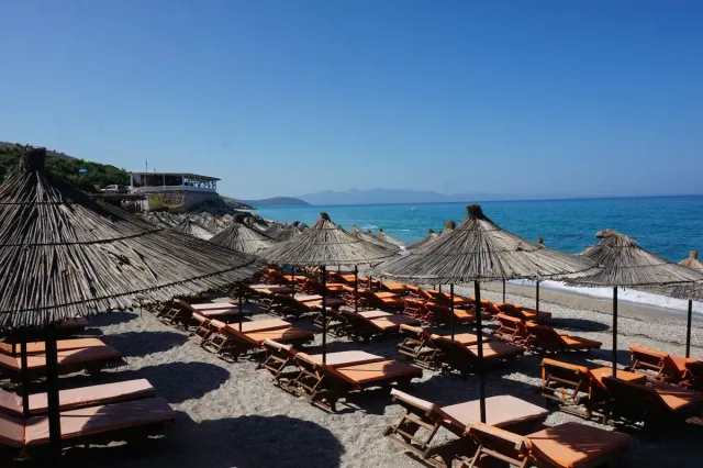 Rows of straw parasols and orange sunbeds on a pebble beach facing a blue sea
