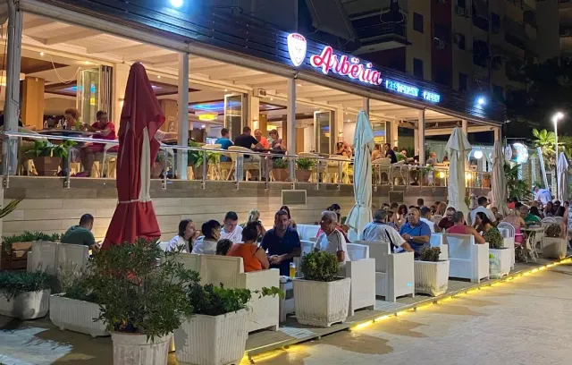 A terrace crowded with diners at night with a lit sign, closed umbrellas and white planters