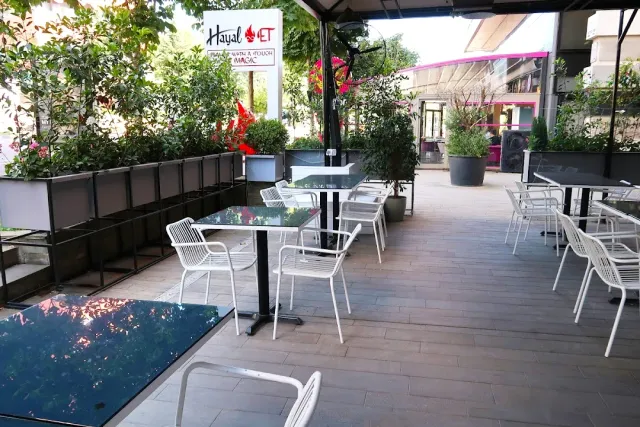 A covered terrace with black tables, white metal chairs and planters of greenery around
