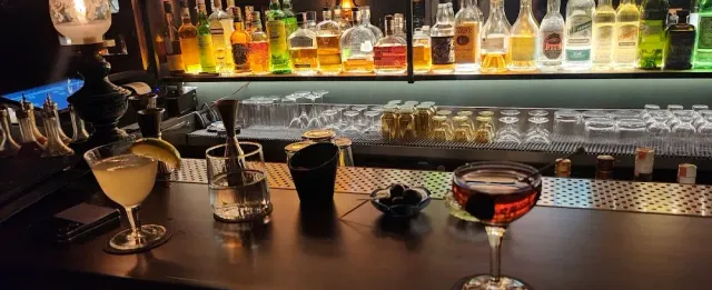 A bar counter at night with two cocktails, a bowl of olives and lit shelves of bottles
