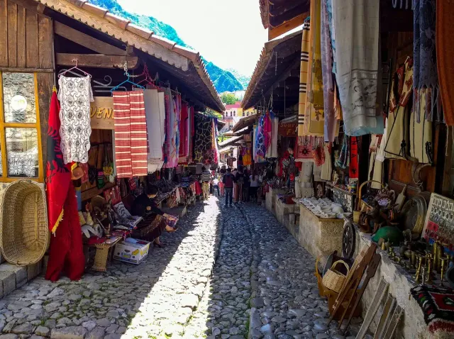 A cobbled lane between wooden shopfronts, with rugs and wares hanging on both sides