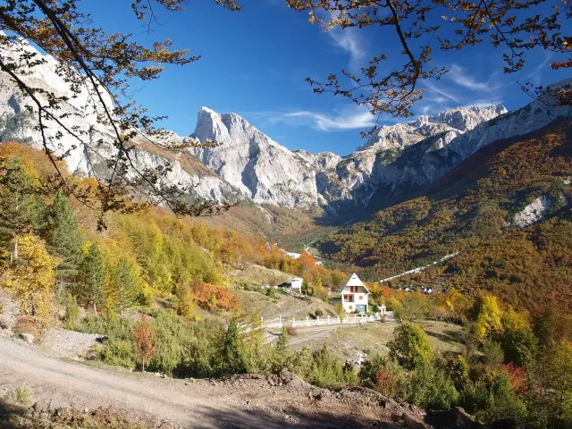 A small stone village with a church in a valley, ringed by autumn forest and snow dusted rock peaks