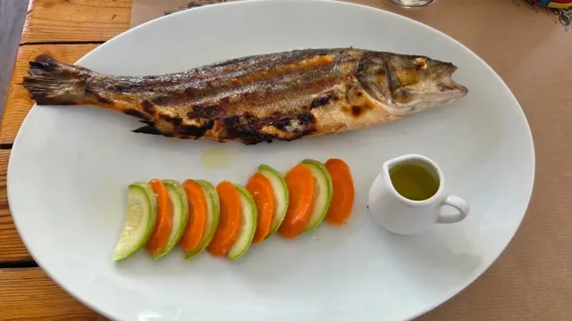 A whole roasted fish on a white plate with zucchini and carrot slices and a jug of olive oil