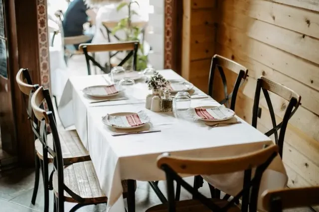 A long table in a white cloth with dark wooden chairs and arranged napkins by a wooden wall