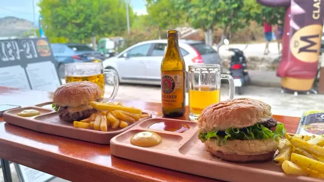 Two burgers on wooden trays with fries, a beer bottle and mugs on an outdoor table