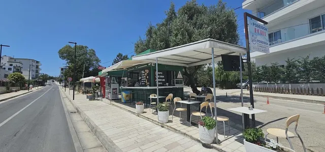 A green kiosk with a white awning and small tables on a wide sidewalk by the road