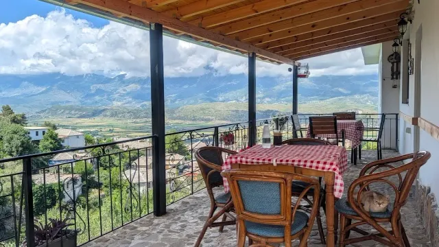A covered balcony with a red checked cloth and cane chairs, a sleeping cat and a view of valley and mountains