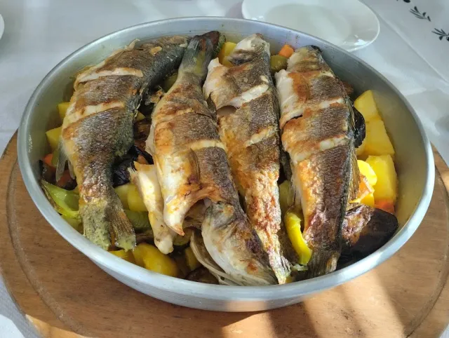 Four whole grilled fish in a metal pan over potatoes, peppers and carrots