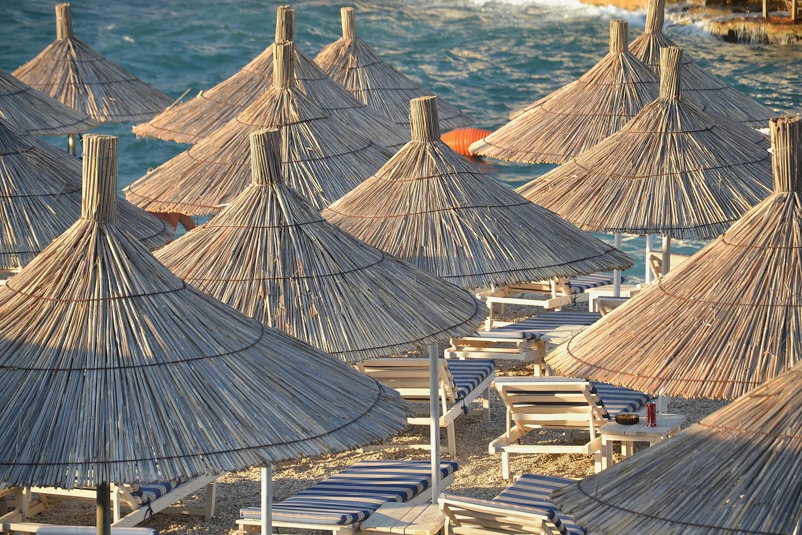 Straw umbrellas and sun loungers lining a pebble beach beside turquoise water in Ksamil